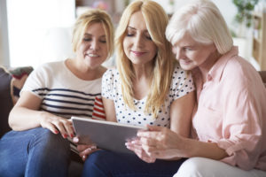 A senior woman looking at a tablet with her two daughters while sitting on a couch