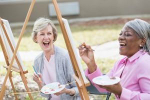 Two senior women laughing as they paint on easels