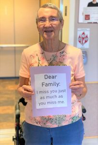 Photo of a long-term care resident holding a sign that says dear family I miss you just as much as you miss me
